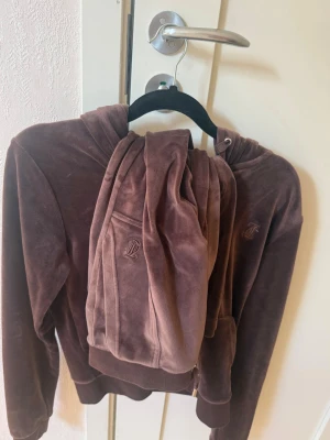 Brunt velourset från Juicy Couture - Mysigt loungewear-set i brun velour från Juicy Couture. Setet består av en hoodie med dragkedja och matchande byxor med resår i midjan. Båda delarna har broderad JC-logga framtill. Perfekt för chill dagar hemma. Använd 5-6 gånger. Tröja storlek S underdel Xs