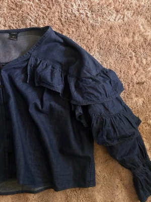 Blå volangblus från Linosei - Trendig blå blus i denim-look från Linosei med långa ärmar och flera volangdetaljer över axlar och ärmar. Blusen har rund halsringning och knäppning framtill. Perfekt för dig som gillar en unik och cool stil.