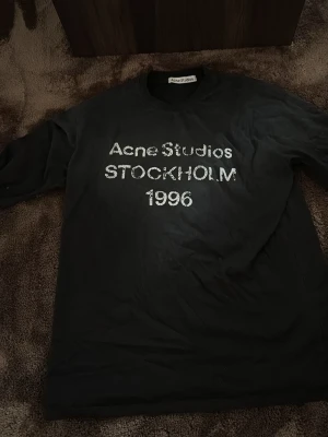 Svart Acne Studios t-shirt - Snygg svart t-shirt från Acne Studios med tryck i vitt framtill där det står 'Acne Studios STOCKHOLM 1996'. Klassisk rund hals och rak passform. Perfekt för dig som gillar stilrena och trendiga plagg.