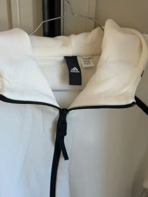 Vit hoodie med dragkedja från Adidas - Snygg vit hoodie från Adidas med svart dragkedja framtill och klassisk huva. Tillverkad i mjukt bomullsmaterial som känns skönt mot huden. Perfekt för en sportig och avslappnad stil. Adidas-logga på insidan av kragen.
