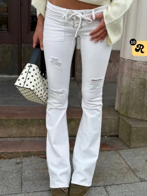 Vita bootcut jeans med snörning - Säljer ett par vita bootcut jeans med snörning i midjan och slitna detaljer på låren och knäna. Jeansen har hög midja och är tillverkade i stretchigt denimtyg för en bekväm passform. Perfekta för en trendig och avslappnad look.