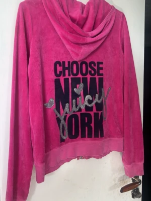 Juicy Couture rosa velour hoodie - Snygg rosa hoodie i mjuk velour från Juicy Couture med dragkedja och fickor fram. Baksidan har stor text 'CHOOSE NEW YORK Juicy' i svart och silverglitter. Perfekt för dig som gillar statement-plagg och bekväma material.