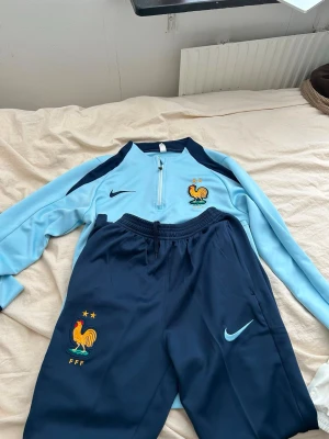 Nike Frankrike träningsset blå - Snyggt träningsset från Nike med Frankrikes landslagslogga. Tröjan är ljusblå med mörkblå detaljer och halv dragkedja, byxorna är mörkblå med broderad tupp och FFF-logga. Perfekt för fotbollsträning eller chill. Materialet är mjukt och stretchigt.