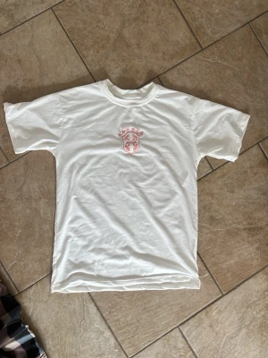 Vit t-shirt med röd logga - Vit t-shirt i bomull med korta ärmar och en röd logga på bröstet. Klassisk rund halsringning och normal passform. Perfekt till jeans eller shorts för en enkel och clean look.