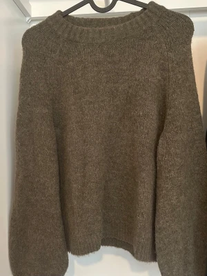 Grön/brun stickad tröja med rund hals - Mysig grön/brun stickad tröja med rund halsringning och breda ärmar. Tröjan har en enkel, oversized passform och ribbstickade muddar vid hals, ärmslut och nederkant. Perfekt för lager på lager under kyliga dagar.