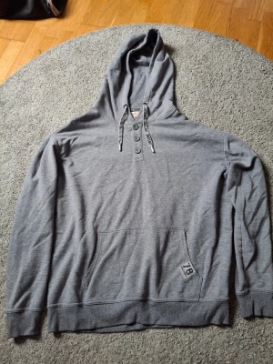Grå hoodie från Gin Tonic - Snygg grå hoodie från Gin Tonic med knappar vid halsen och stor huva. Tröjan har magficka och en liten patch med siffran 78 nedtill. Tillverkad i mjukt bomullsmaterial, perfekt för en avslappnad stil.