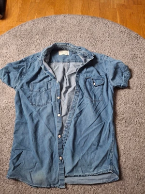 Blå jeansskjorta från Jack & Jones - Säljer en klassisk blå jeansskjorta från Jack & Jones med korta ärmar och två bröstfickor. Skjortan har knappar framtill och är tillverkad i mjukt bomullsmaterial. Perfekt för en avslappnad och cool stil.