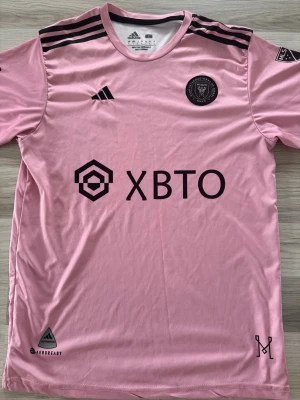 Inter Miami Messi matchtröja rosa - Säljer en rosa Inter Miami CF matchtröja från Adidas med Messi och nummer 10 på ryggen. Tröjan har svarta detaljer, klubbmärke på bröstet, sponsortryck och AEROREADY-material som andas. Perfekt för fotbollsfans som vill sticka ut på läktaren.