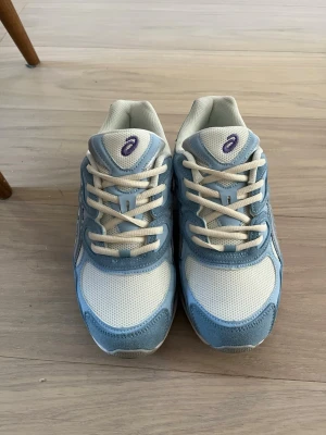 ASICS blåvita sneakers - Säljer ett par snygga sneakers från ASICS i ljusblått och vitt med mesh och mockadetaljer. Skorna har klassiska låga snitt och vita snören. Perfekta för dig som gillar sportig stil och vill ha bekväma skor med cool design.