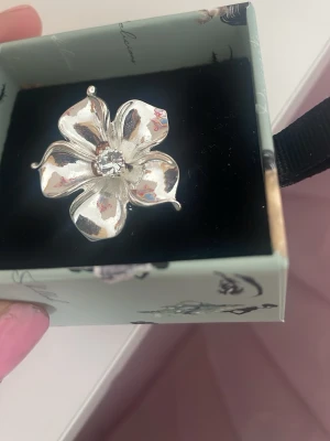 Blommig silverring med kristall - Söt blomring i silver från märket ioaku. Den är justerbar och lätt att använda. Kommer inte till användning längre och den har en lätt defekt nära mitten. Titta på bilden. Ask medföljer! 