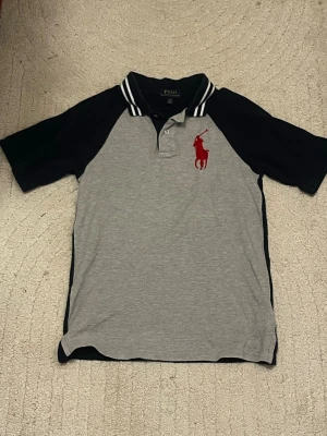 Grå och marinblå piké från Polo Ralph Lauren - Snygg pikétröja från Polo Ralph Lauren i grått och marinblått med klassisk krage och vita ränder. Stor röd logga på bröstet och tryck på ryggen med texten 'Polo RL Athletics'. Perfekt för en sportig och avslappnad stil.