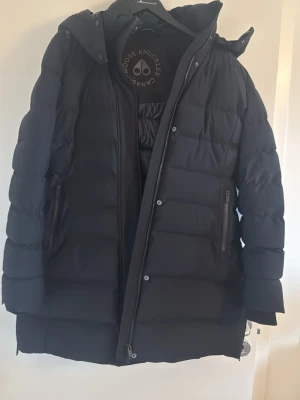 Svart dunjacka Moose Knuckles - Snygg svart dunjacka från Moose Knuckles med lång passform och huva. Jackan har dragkedja framtill, två sidofickor med dragkedja och diskret logga på insidan. Perfekt för kalla vinterdagar och riktigt varm. XL men passar L