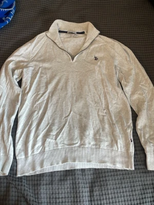 Ljusgrå tröja från U.S. Polo Assn. - Ljusgrå långärmad tröja från U.S. Polo Assn. med halvzip och klassisk krage. Diskret logga broderad på bröstet och ribbade muddar vid ärmslut och nederkant. Perfekt för en avslappnad och stilren look. Tröjan är mer s än m i verkligheten obs.