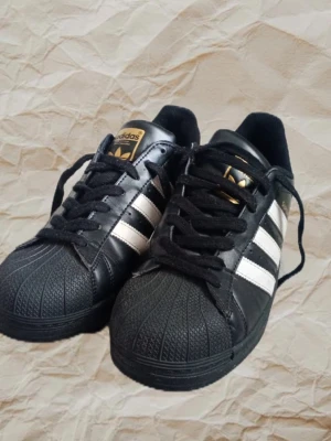 Adidas Superstar svarta sneakers - Klassiska Adidas Superstar sneakers i svart läder med vita ränder och gulddetaljer. Skorna har den ikoniska snäckformade tån och svart sula. Låg modell med svart snörning och vit hälkappa med Adidas-logga. Perfekta för en streetwear-look.
