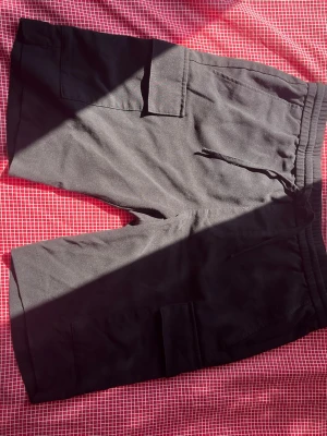 Svarta cargo shorts med snörning - Säljer ett par svarta cargo shorts med snörning i midjan och flera praktiska fickor på sidorna. Shortsen har en loose passform och är perfekta för chill dagar. Tillverkade i mjukt bomullsmaterial för extra komfort.