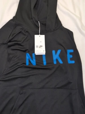 Svart linne med luva  från Nike med blå text smal  - Svart linne med luva  från Nike med stor blå NIKE-text på bröstet. Klassisk huva och stor magficka framtill. Tillverkad i mjukt material som känns skönt mot huden. Perfekt för en avslappnad och sportig stil helt ny passar medium med 