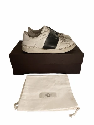 Valentino Garavani vita sneakers - Snygga vita sneakers från Valentino Garavani med bred svart läderdetalj över mitten och klassiska nitar på sulan. Skorna har rund tå, vita snören och är tillverkade i läder. Nypris :4900 säljer endast för 999