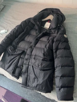 Svart dunjacka från Moncler - Säljer en svart dunjacka från Moncler med klassisk puffer-design och stora fickor framtill. Jackan har huva, dragkedja och knappar samt Moncler-logga på ärmen. Perfekt för kalla dagar och riktigt snygg att bära till jeans eller joggers.