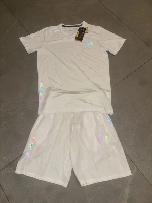 Vitt set med holografiska detaljer - Säljer ett vitt tvådelat set med t-shirt och shorts. Setet har holografiska loggor och detaljer på både tröjan och shortsen. Materialet ser ut att vara mjukt och bekvämt, perfekt för en avslappnad stil. Passar dig som gillar en clean look med lite extra bling.