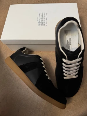 Maison Margiela Gats - Svarta sneakers från Maison Margiela med klassisk design, vita snören och beige gummisula. Skorna har en mix av läder och mocka på ovandelen och en låg silhuett. Perfekta för dig som gillar stilrena och tidlösa sneakers med en lyxig känsla. Helt nya och oanvända 