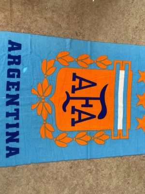 Argentina blå-orange sovpläd - Mysig sovpläd i ljusblått med stort orange tryck av Argentinas emblem och texten 'ARGENTINA' längs sidan. Pläden har dekorativa detaljer i blått, vitt och orange samt tre stjärnor. Perfekt för att chilla hemma eller ta med på resan.