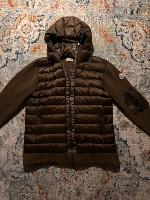 Moncler cardigan  - Snygg dunväst från Moncler med stickade ärmar och huva. Västen har två fickor med dragkedja framtill och en Moncler-logga på ärmen. Materialet är en mix av dun och stickat tyg, perfekt för kyliga dagar.