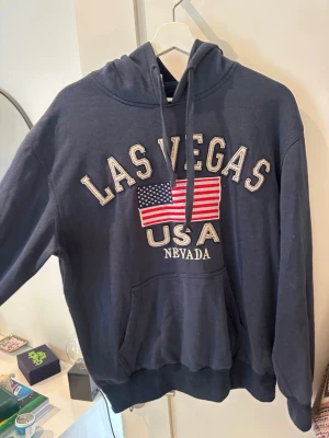 Mörkblå Las Vegas hoodie med tryck - Mörkblå hoodie med huva och känguruficka. Framtill står det 'Las Vegas USA Nevada' och en stor amerikansk flagga är tryckt mitt på bröstet. 