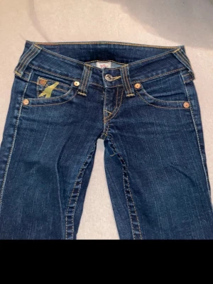 Mörkblå bootcut jeans från true religion - Snygga mörkblå bootcut jeans från true religion med klassiska kontrastsömmar och fem fickor. Jeansen har en gul detalj på myntfickan och är tillverkade i slitstark denim. Perfekta för dig som gillar en tidlös och avslappnad stil.