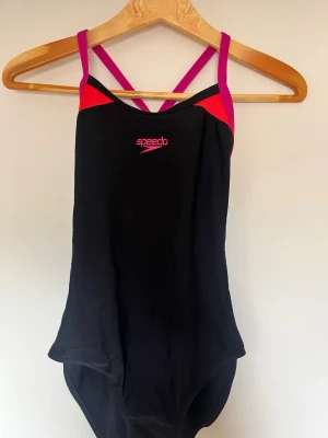 Svart baddräkt från Speedo - Snygg svart baddräkt från Speedo med rosa och röda detaljer samt tunna axelband som korsas i ryggen. Tillverkad i ett stretchigt material som passar perfekt för simning och träning. Klassisk sportig design med Speedo-logga framtill. Använd 2 gånger 
