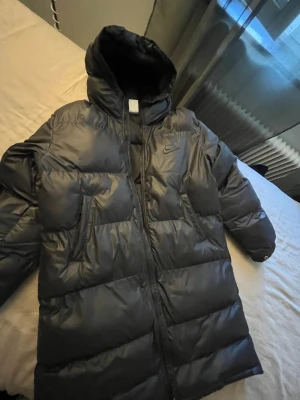 Svart lång pufferjacka från Nike - Säljer en svart lång pufferjacka från Nike med huva och dragkedja framtill. Jackan har en blank finish och klassisk Nike-logga på bröstet. Perfekt för kalla vinterdagar och riktigt skön att bära. Materialet är syntet och jackan har en oversized look.