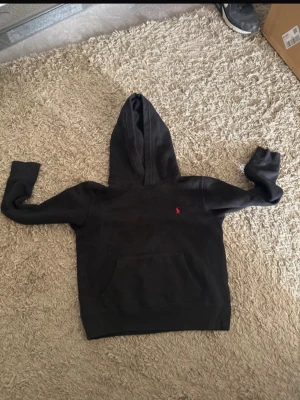 Svart hoodie från Polo Ralph Lauren Barn M inte vuxen  - Svart hoodie från Polo Ralph Lauren med klassisk huva och känguruficka framtill. Liten röd logga broderad på bröstet. Mjuk bomullskänsla och ribbade muddar vid ärmslut och nederkant. Perfekt för en chill och avslappnad stil.