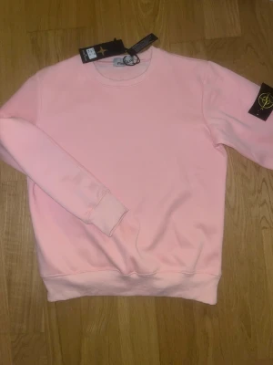 Ljusrosa sweatshirt från Stone Island - Ljusrosa sweatshirt från Stone Island med klassisk rund hals och ribbade muddar. Tröjan har den ikoniska loggan på vänster ärm och är tillverkad i mjukt bomullsmaterial. Perfekt för en avslappnad och trendig look.