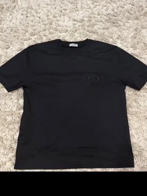 Svart t-shirt från Dior med diskret logga - Snygg svart t-shirt från Dior med en diskret broderad CD-logga på bröstet. Klassisk passform och rund halsringning. Tillverkad i mjuk bomull som känns skön mot huden. Perfekt basplagg för en clean och stilren look.