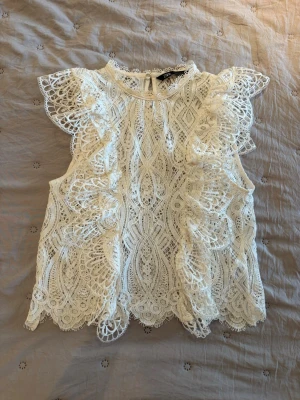 Vit spetsblus från Zara - Supersöt vit spetsblus från Zara med detaljerade broderier och volang vid axlarna. Blusen är ärmlös och har rund halsringning samt knapp i nacken. Perfekt för en chic och romantisk look.