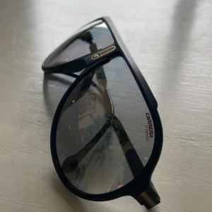 Svarta Carrera pilotbågar solglasögon - Snygga svarta solglasögon från Carrera med klassisk pilotform och gråtonade linser. Ramen är i matt svart plast med guldiga Carrera-detaljer på både front och skalmar. Perfekt accessoar för en cool och sportig look.