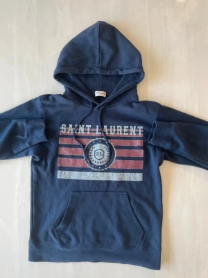 Mörkblå Saint Laurent hoodie - Mörkblå hoodie från Saint Laurent med tryck framtill i vitt och rött, samt texten 'SAINT LAURENT'. Klassisk känguruficka och justerbar huva med snören. Tillverkad i mjukt bomullsmaterial, perfekt för en avslappnad streetwear-look. Storlek S sitter som Xs, helt ny  kan byta emot M