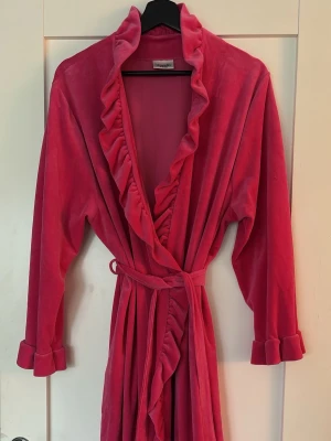 Rosa volangmorgonrock från Damella - Mysig rosa morgonrock från Damella i mjuk velour. Knälång modell med långa ärmar, volangkant längs hela öppningen och nederkanten samt ett matchande midjeband. Storlek saknas men gissar på L.