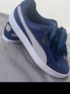 Blå Puma Basket sneakers med sidenband - Snygga blå Puma Basket sneakers med vit sula och vit sidostripes. Skorna har breda sidenband som skosnören och gulddetaljer vid snörningen. Ovandelen är i mocka med diskret prickmönster och insidan har ett grafiskt mönster.