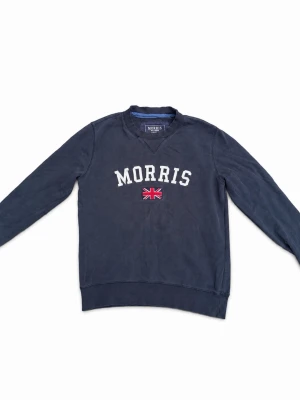 Marinblå sweatshirt från Morris - Klassisk marinblå sweatshirt från Morris med vit broderad logga och den klassiska brittiska flaggan på bröstet. Tröjan har rund halsringning, ribbade muddar och en patch på ärmen. Är en liten äldre verision som inte längre är tillgänglig i butik eller hemsida, nu hos oss för endast 300kr och priset är självklart diskuterbart! Kontakta oss vid frågor och funderingar!