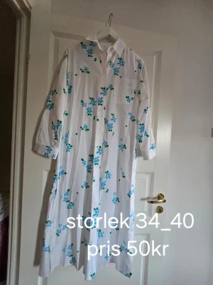 Vit blommig långklänning - Långklänning i vitt med blå och gröna blommor. Klänningen har krage, knappar framtill och trekvartsärm. Perfekt för sommarens varma dagar och har en avslappnad passform.