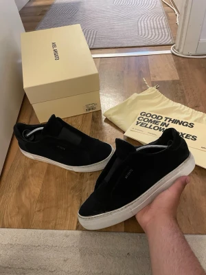 Svarta sneakers från Axel Arigato - Stilrena svarta sneakers från Axel Arigato i mocka med vit tjock sula. Diskret logga på sidan och minimalistisk design utan snörning. Perfekta för dig som gillar en clean look och vill ha något som funkar till det mesta. Box medföljer ej och dustbag.bix 
