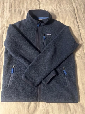 Blå fleecejacka från Patagonia - Mysig blå fleecejacka från Patagonia med hel dragkedja framtill och tre fickor med blå detaljer på dragkedjorna. Jackan har hög krage och är tillverkad i mjukt, värmande fleecematerial. Perfekt för kyliga dagar och outdoor-häng.