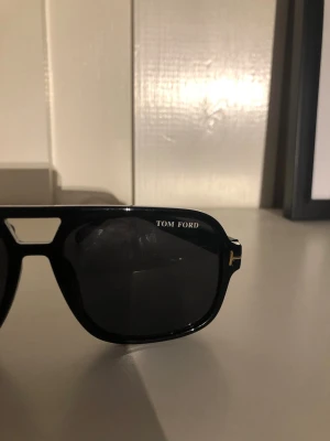 Svarta Tom Ford solglasögon - Snygga svarta solglasögon från Tom Ford med klassisk pilotform och mörka glas. Bågarna är i blank plast med guldfärgade T-detaljer vid gångjärnen. Perfekta för dig som vill ha en stilren och exklusiv look.