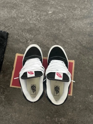 Svarta Vans Old Skool sneakers - Klassiska Vans Old Skool sneakers i svart mocka och canvas med vit sula och vit sidostrip. Skorna har vita skosnören och den ikoniska röda Vans-loggan på plösen. Perfekta för en avslappnad streetstil.