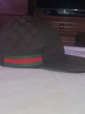 Gucci keps med GG-mönster - Säljer en snygg keps från Gucci i mörkbrunt tyg med klassiskt GG-mönster över hela kepsen. På sidan finns en grön och röd rand som ger en ikonisk touch. Perfekt för dig som vill ha en exklusiv och trendig accessoar.