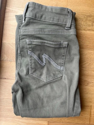 Trendiga khakijeans i fint skick - Snygga khakifärgade jeans med dekor på bakfickan! Superfint skick, inga fläckar men dom är lite slitna nere vid fötterna men inget som syns Sitter jättefint och passar till allt. Normal midja, stretchigt material och sköna att bära. Storlek:34 Nypris ca 600kr 