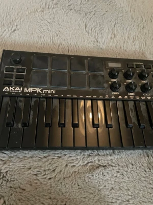AKAI MPK mini MIDI-kontroller - Säljer en svart AKAI MPK mini MIDI-kontroller med 25 tangenter, åtta pads och flera rattar för att styra ljud och effekter. Perfekt för musikproduktion och beatmaking. Kompakt design i plast, smidig att ta med till studion eller hemmet.
