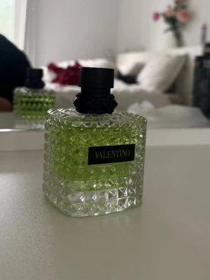 Valentino parfym grön glasflaska - Säljer en snygg parfym från Valentino i en fyrkantig glasflaska med nitar och coolt mönster. Flaskan har en grön tonad vätska och svart kork med detaljer. Perfekt för dig som gillar exklusiva dofter och stilren design.