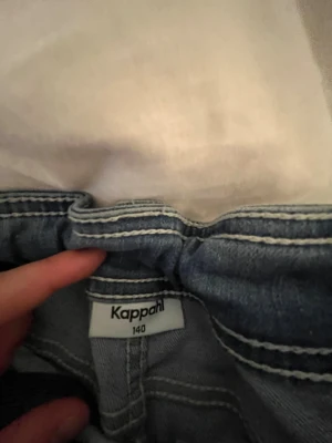 Blå denim jumpsuit från Kappahl - Säljer en blå denim jumpsuit från Kappahl i storlek 140. Jumpsuiten har klassiska jeansdetaljer med synliga sömmar, fickor och silverfärgade knappar framtill. Perfekt för dig som gillar en avslappnad och cool stil.