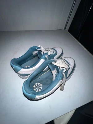 Nike Turkosa Vita storlek 39 - Snygga sneakers i vitt och turkost med klassisk låg siluett. Skorna har perforerad tå, vita snören och en kontrasterande turkos detalj längs sidan och hälen. Insidan är fodrad i turkost tyg och yttersulan är vit med grå kant.